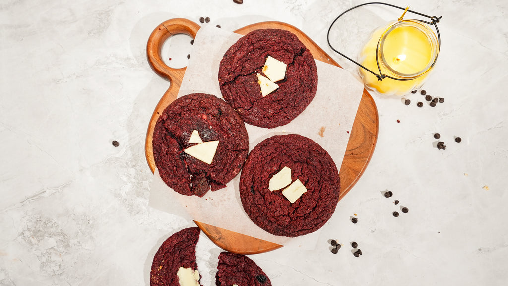 Three red velvet cookies topped with white chocolate pieces, arranged on a wooden board beside a lit candle.