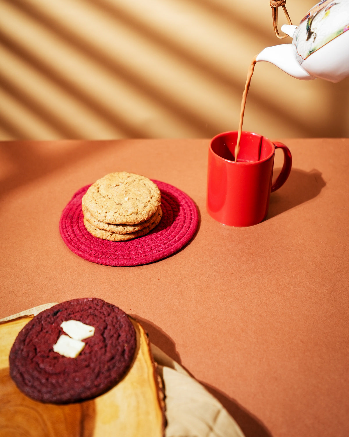 A stack of Chai Latte cookies on a red mat, beside chai being poured into a red mug from a floral teapot.
