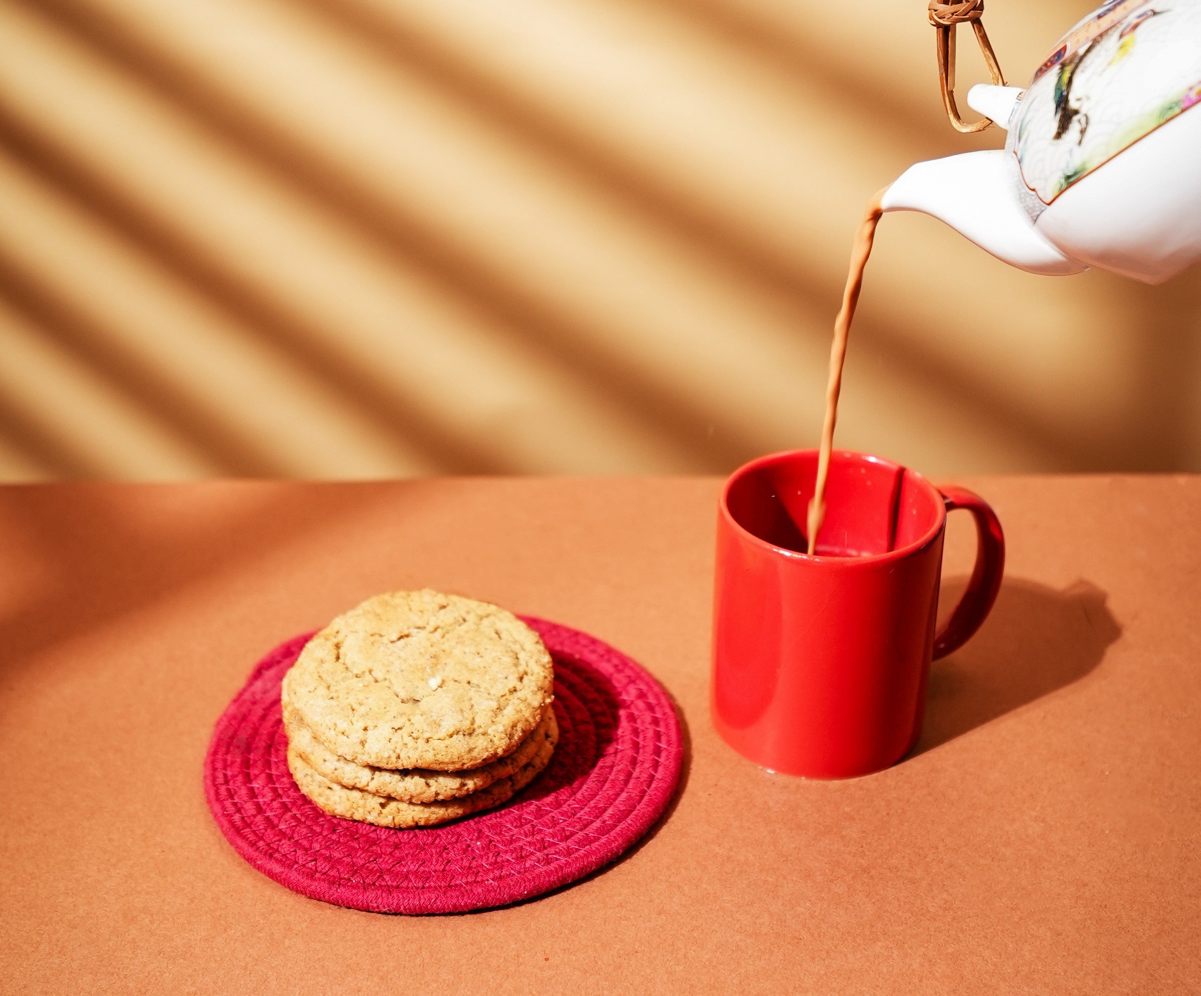 A stack of three chai spiced cookies beside a red mug, with chai being poured from a teapot.