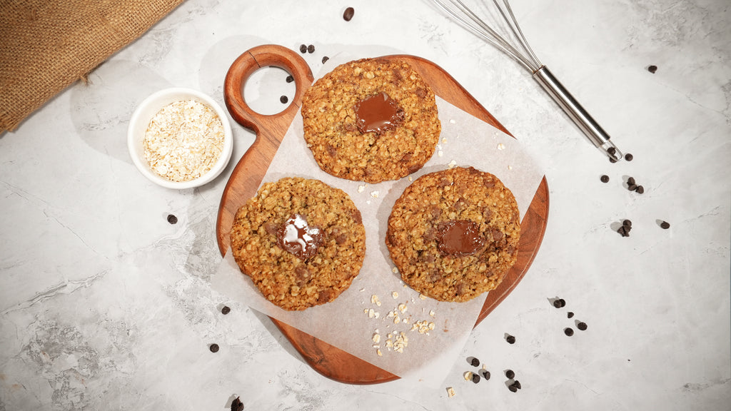 Three oat cookies with melted dark chocolate centres on a wooden board, beside a bowl of rolled oats.